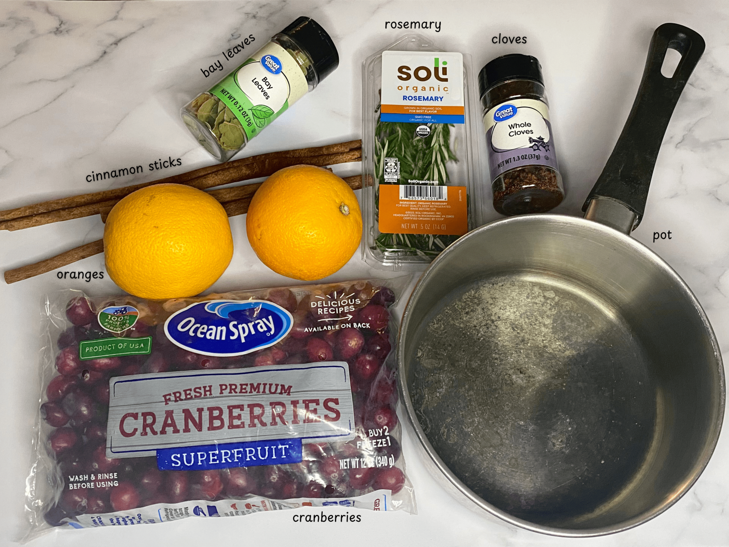 rosemary, cinnamon sticks, oranges, bay leave, cloves, cranberries, and a pot on top of a marble countertop