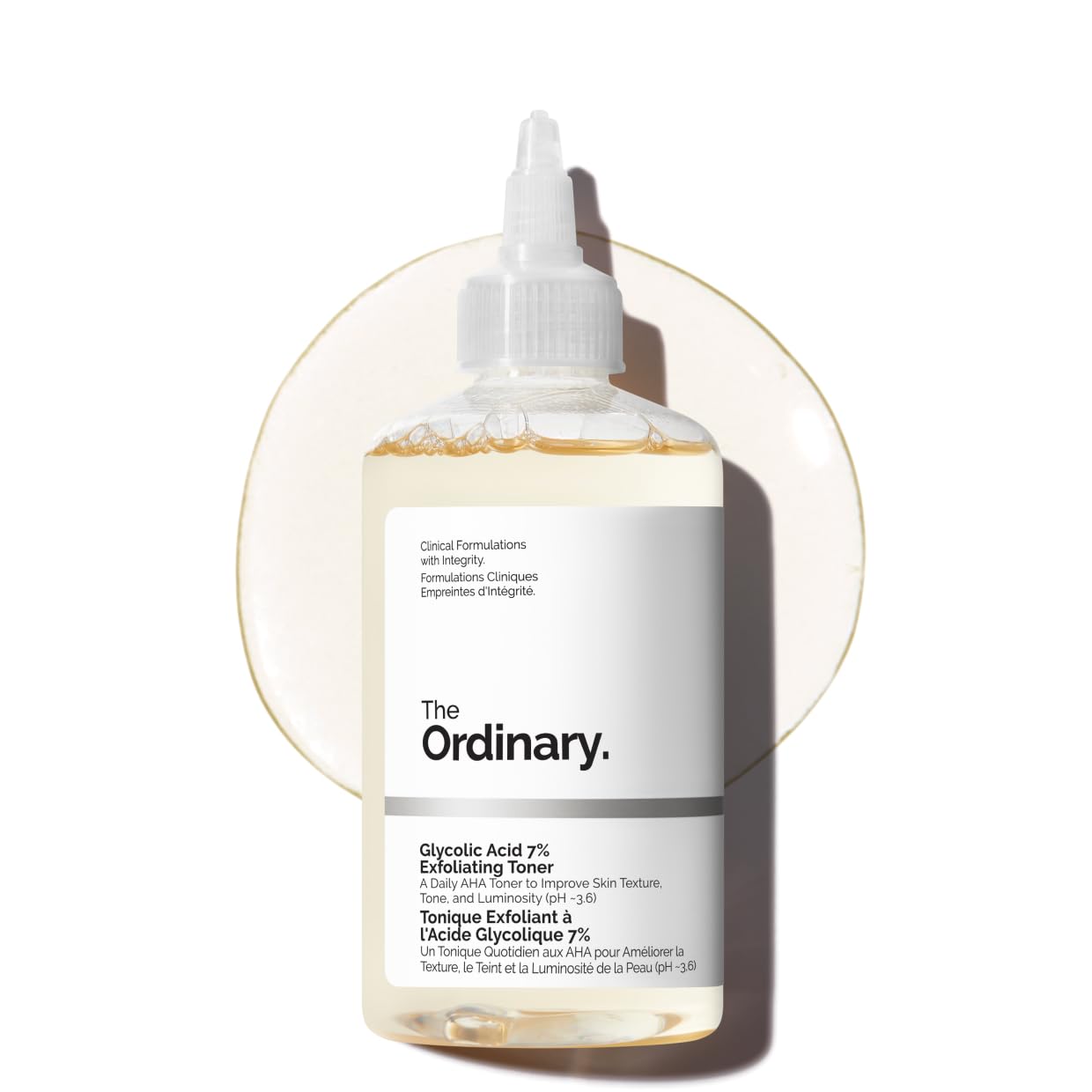 A clear bottle of The Ordinary Glycolic Acid 7% Exfoliating Toner lying flat against a white background with a circular smear of the liquid product visible behind it.