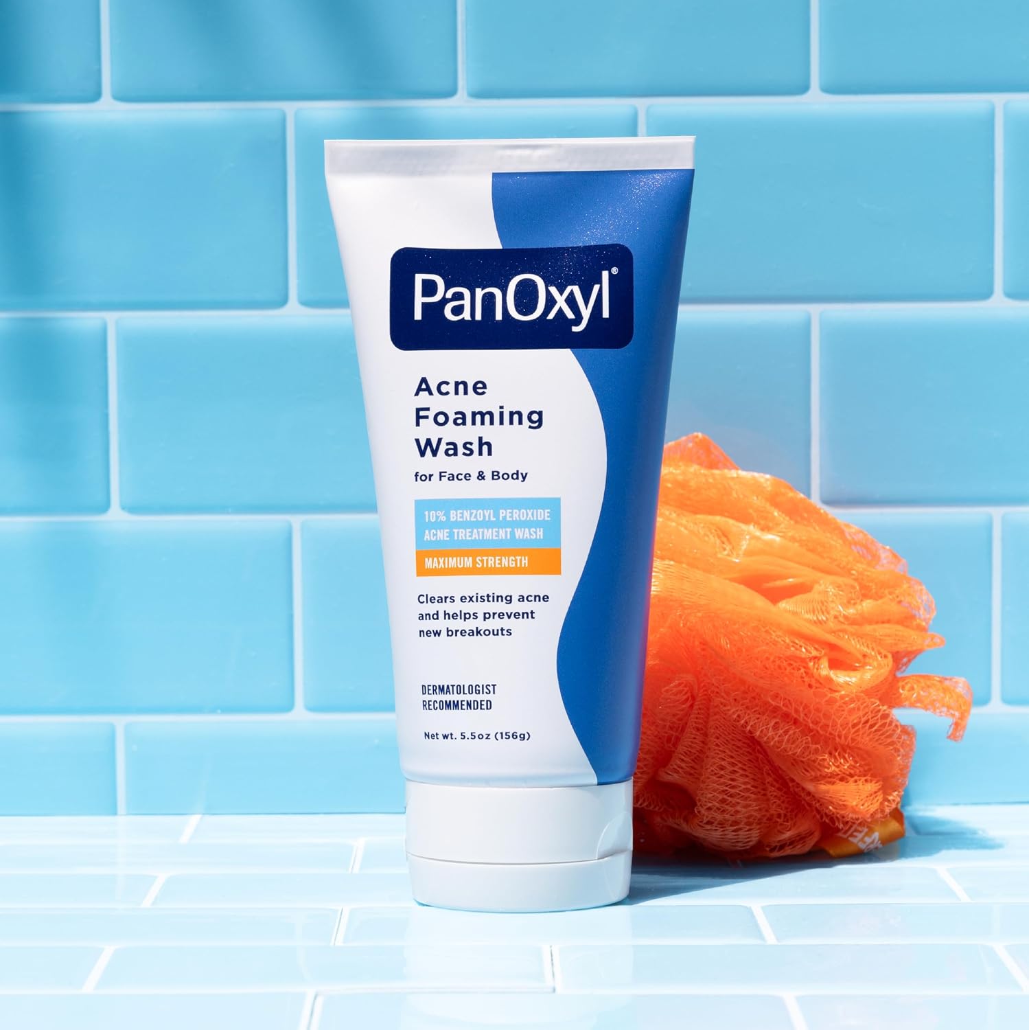 A blue and white tube of PanOxyl Acne Foaming Wash with 10% Benzoyl Peroxide sitting on light blue bathroom tiles next to a bright orange shower loofah.